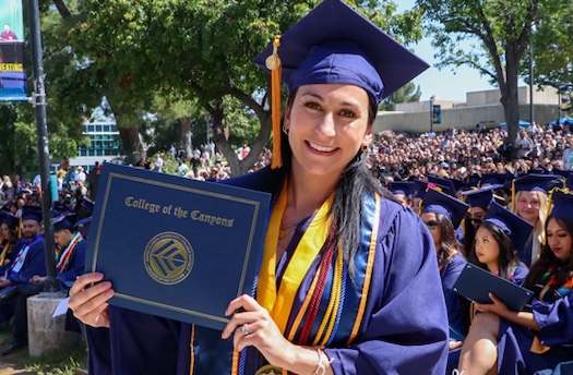 A female student in cap and gown smiles while holding up her degree during the commencement ceremony in the Honor Grove. Fellow graduates are seated behind her.