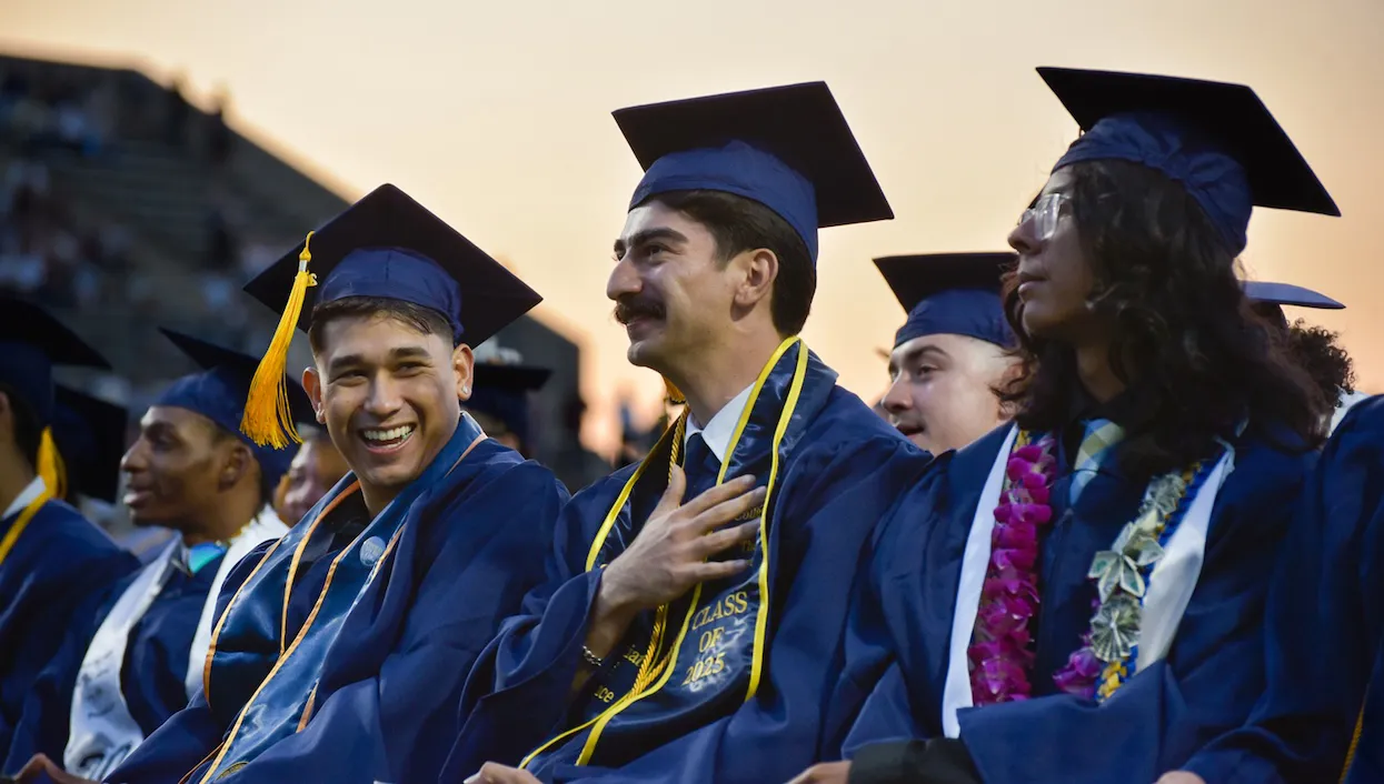 Graduates in blue caps and gowns sit in a row at an outdoor commencement ceremony, smiling and looking toward the stage at sunset.