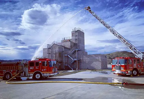 Fire trucks from the LA County Fire Department display their firefighting capabilities with hoses spraying water at a training facility.