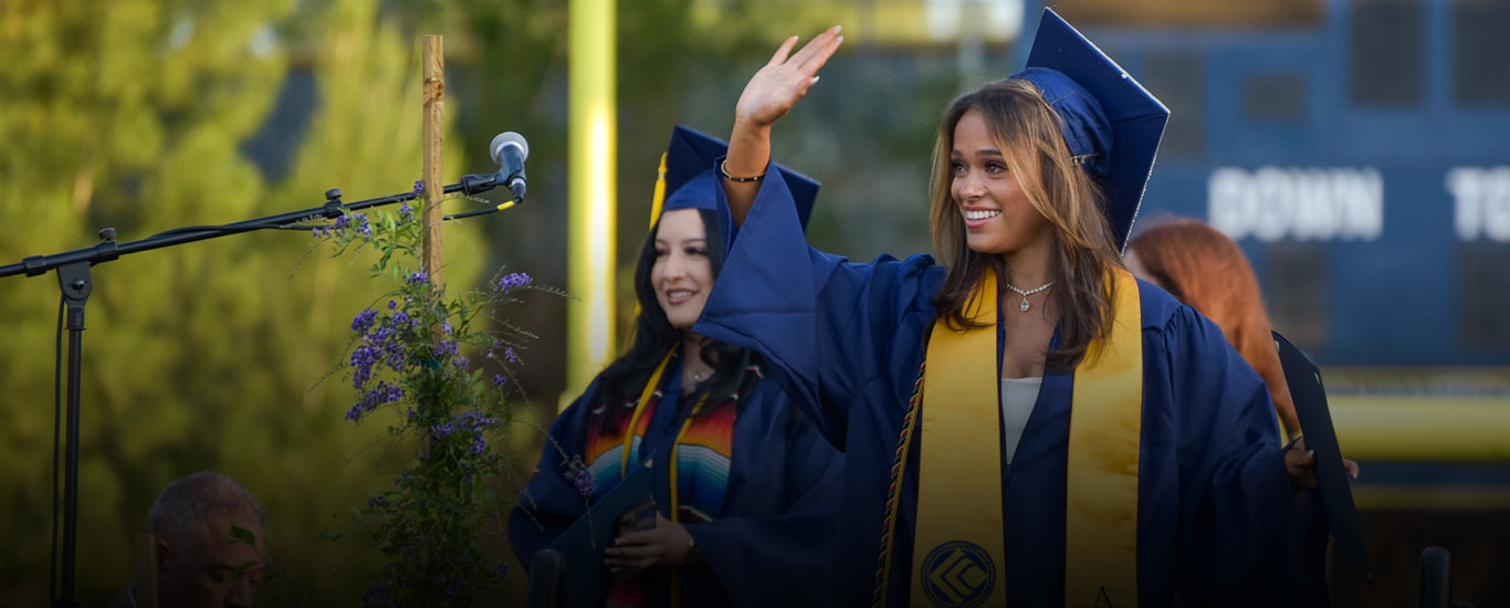 A female graduate in cap and gown smiles and waves as she picks up her diploma.