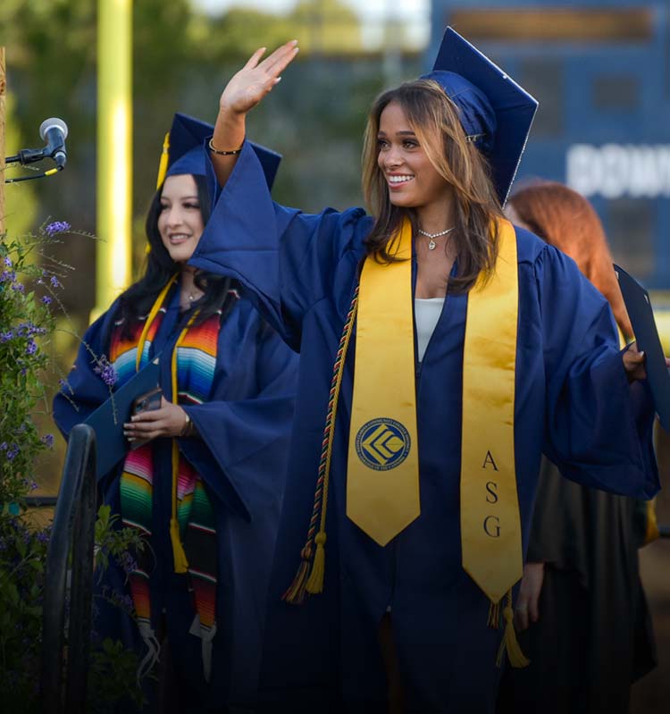 A female graduate in cap and gown smiles and waves as she picks up her diploma.