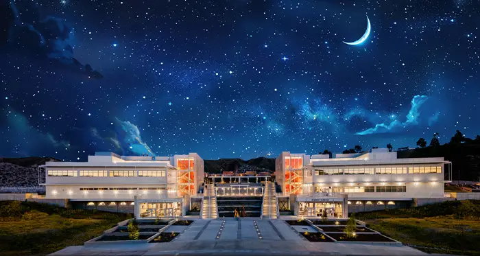 Modern, brightly lit campus buildings at night beneath a star-filled sky with a crescent moon, viewed symmetrically from a central walkway.