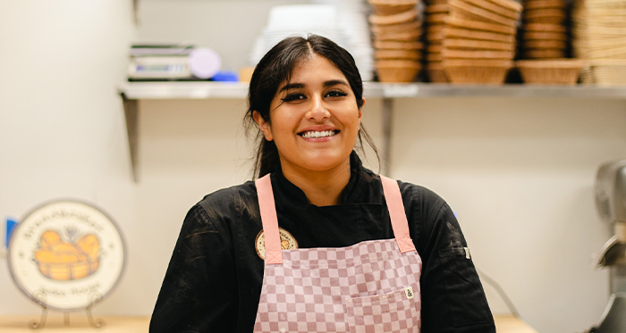 A smiling Leslie Navarro, wearing a pink checkered apron, stands in her bakery kitchen with shelves of baskets in the background.
