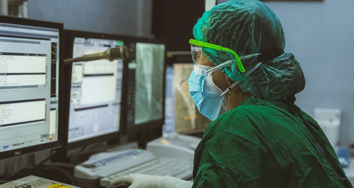 A nurse in scrubs and protective gear peers at data on a computer screen.