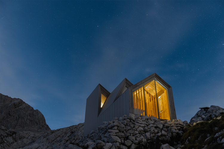 Modern home at night, with peaked roof, lit windows aglow.