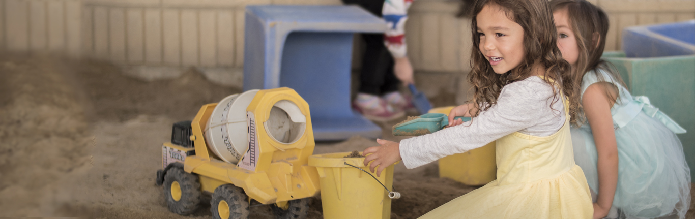 Little girls in dresses playing with truck in the sandbox at the Early Childhood Education program. 